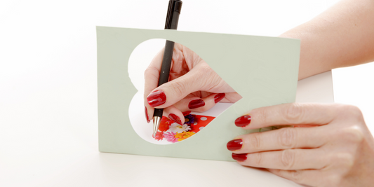 A female hand with red painted nails writing a card with a heart shaped cut out of the front with a white background